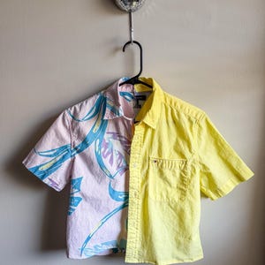 May include: A short-sleeved shirt, split vertically with a floral pattern on one side and solid yellow on the other. The shirt hangs on a black hanger. The floral side is pastel pink with blue and purple leaves.
