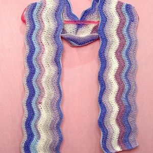 May include: A crochet scarf featuring a wavy ripple pattern in alternating horizontal stripes of white, light blue, medium blue, and lavender. The scarf is draped over a bright pink plastic hanger, showcasing its textured yarn construction.