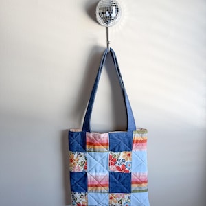 May include: A quilted tote bag with a patchwork design. The bag features squares of denim, floral, and striped fabrics in shades of blue, pink, orange, and green. The tote has denim handles and is hanging from a silver disco ball hook.