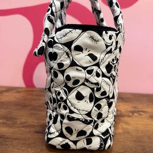 May include: A small, quilted tote bag featuring a repeating pattern of Jack Skellington faces in black and white. The bag has two padded handles and a black interior lining. The bag is sitting on a wooden surface.