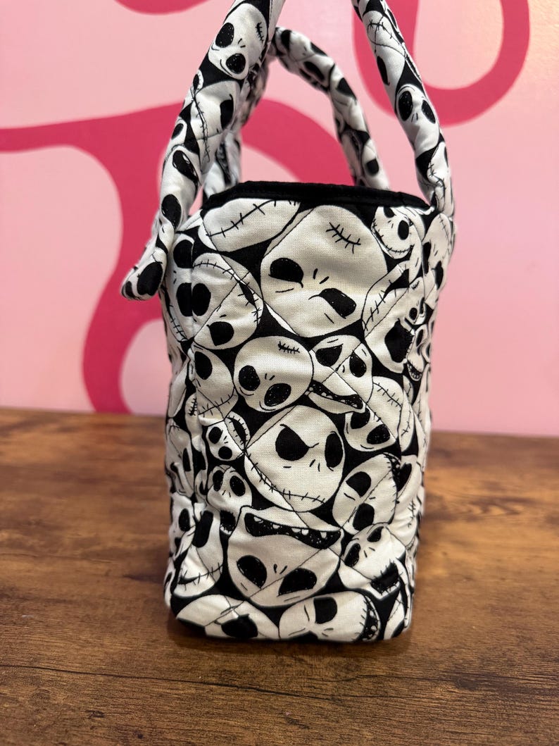 May include: A small, quilted tote bag featuring a repeating pattern of Jack Skellington faces in black and white. The bag has two padded handles and a black interior lining. The bag is sitting on a wooden surface.