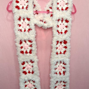 May include: A long, handmade crochet scarf constructed from square motifs. Each square features a light pink center, surrounded by white and bright red yarn. The entire scarf is edged with a fuzzy, white, faux-fur style yarn texture.