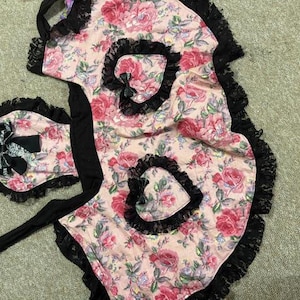 Puede incluir: Un delantal floral rosa y un gorro a juego con ribete de encaje negro. El delantal presenta un diseño en forma de corazón y un lazo negro. El estampado floral incluye rosas rojas y hojas verdes.