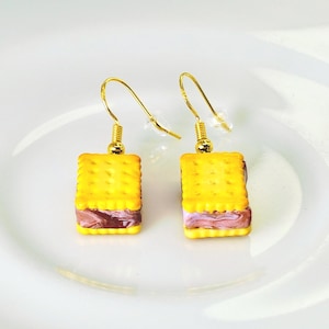 May include: A pair of novelty earrings shaped like yellow cracker sandwiches with a purple and brown filling. The earrings have gold-colored hooks and are displayed on a white plate.