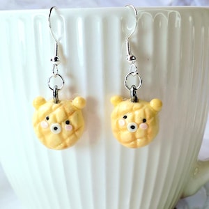 Puede incluir: Un par de pendientes colgantes plateados. Cada pendiente presenta un pequeño dije amarillo con forma de oso con una superficie texturizada, ojos negros y mejillas rosadas. Los pendientes se muestran sobre un fondo blanco.
