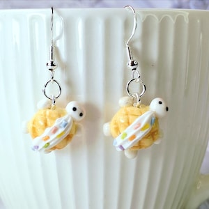 Op de afbeelding: Een paar zilverkleurige oorhangers met gele schildpadbedels, witte koppen en kleurrijke surfplanken. De oorbellen worden gepresenteerd tegen een witte achtergrond, waarbij de details van de sieraden worden benadrukt.