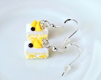 Pode incluir: Brincos prateados com fatias de bolo em miniatura. Cada fatia tem uma camada superior branca, recheio amarelo e é coberta com fruta amarela e uma baga escura. Em uma superfície branca.