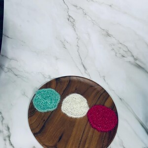 Puede incluir: Un plato de madera contiene tres estropajos de ganchillo en verde azulado, blanco y rojo. Los estropajos son redondos y esponjosos. El plato tiene un acabado de vetas de madera natural. Los estropajos miden unos 7 cm de diámetro.