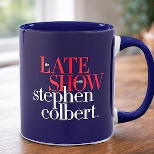 May include: A navy blue ceramic mug with a white interior and handle. The mug features the text "The Late Show with Stephen Colbert" in red and white lettering. The mug is on a wooden surface.