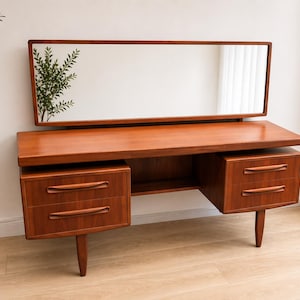 May include: A mid-century modern wooden dressing table with a rectangular mirror. The table has a warm, reddish-brown finish, two sets of drawers, and tapered legs. The mirror is framed in matching wood.