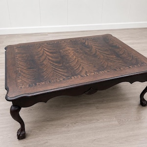 May include: A dark brown, rectangular coffee table with a patterned top and ornate, curved legs. The table has a decorative edge and is made of wood. The legs are claw-footed, adding to the antique aesthetic.