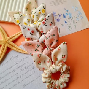 May include: A set of three fabric hair scrunchies featuring tied bow accents. The scrunchies are made of cream and light pink fabric with small floral patterns in red, green, and yellow. Each scrunchie has two upright fabric loops resembling bunny ears.