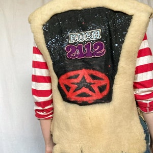 May include: A cream-colored shearling vest with a black denim back panel. The back panel features a red and black star with a black star inside, and the text "RUSH 2112" in purple and pink.