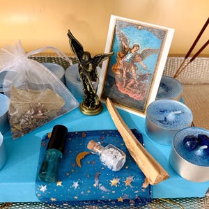 May include: A collection of spiritual items on a blue box, including blue candles, a bronze angel figurine, a small bottle, a card with an angel illustration, incense sticks, and a small bag of herbs. The items are arranged on a silver tray.