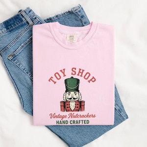 May include: A light pink t-shirt featuring a graphic of a nutcracker in a green hat and red uniform. The text reads TOY SHOP in red, Vintage Nutcrackers in red, and HAND CRAFTED in green. The shirt is paired with blue denim jeans.