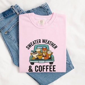 Pode incluir: Uma t-shirt rosa claro com um gráfico de uma carrinha pick-up azul-petróleo carregando chávenas de café e uma raposa com um cachecol. O texto "SWEATER WEATHER & COFFEE" está escrito em letras pretas. A t-shirt está dobrada sobre calças de ganga.