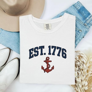 May include: White t-shirt with a navy blue arched "EST. 1776" text and a red anchor graphic. The shirt is folded and displayed with a hat, jeans, and white boots.