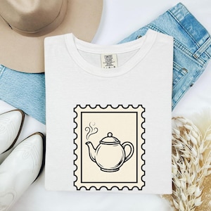 Könnte beinhalten: Weißes Comfort Colors T-Shirt mit einem schwarzen und cremefarbenen Stempeldesign einer Teekanne mit aufsteigendem Dampf. Das Shirt ist auf einer hellen Oberfläche gefaltet, mit blauen Jeans und weißen Stiefeln.