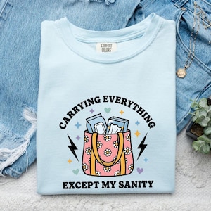 May include: A light blue t-shirt featuring a graphic of a pink tote bag with a white daisy pattern, filled with blue boxes. The text reads CARRYING EVERYTHING EXCEPT MY SANITY, accented by small colourful stars, lightning bolts, and heart icons.