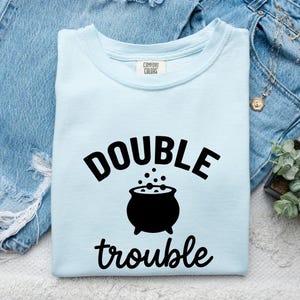 Pode incluir: Uma t-shirt azul-clara com decote redondo e uma impressão gráfica preta na frente. O design apresenta o texto "DOUBLE" acima de um ícone de caldeirão preto com bolhas a subir, e a palavra "trouble" escrita em cursivo abaixo do caldeirão.