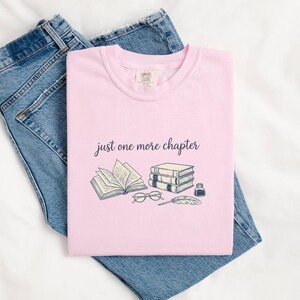 May include: A light pink t-shirt with the text "just one more chapter" and a graphic of books, glasses, a feather, and ink. The shirt is laid on top of a pair of blue jeans.