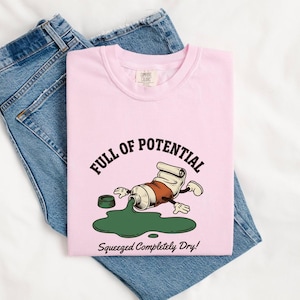 Op de afbeelding: Een lichtroze t-shirt met een afbeelding van een cartoon verftube met armen en benen, die groene verf morst. De tekst luidt "FULL OF POTENTIAL" in een boog erboven en "Squeezed Completely Dry!" eronder. Gecombineerd met een blauwe spijkerbroek.