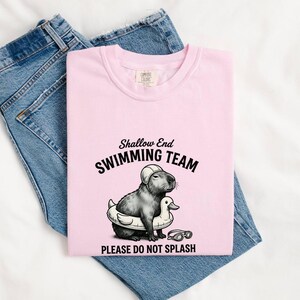 Peut inclure: T-shirt rose clair avec l'inscription "Shallow End Swimming Team" et un dessin d'un capybara avec un bonnet de bain et une bouée canard. Le t-shirt est posé sur un jean bleu.