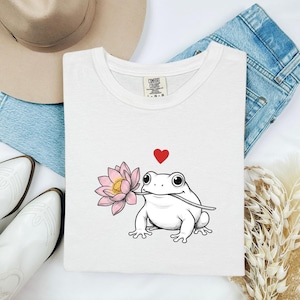 May include: White t-shirt featuring a cartoon frog holding a pink lotus flower in its mouth, with a red heart above it. The shirt has a crew neck and a Comfort Colors tag. The design is centered on the front of the shirt.