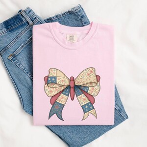 May include: A light pink t-shirt featuring a large bow design. The bow has a patchwork pattern with floral and star details in shades of pink, blue, and cream. The shirt is laid on top of a pair of blue denim jeans.