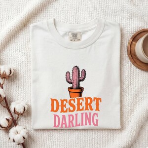 May include: White t-shirt featuring a pink cactus in a terracotta pot, with the words "DESERT DARLING" in orange and pink. The shirt is folded on a white surface. The t-shirt has a crew neck.