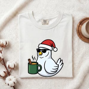 May include: A white t-shirt featuring a graphic of a white bird wearing a red and white Santa hat and black sunglasses. The bird is holding a green mug containing a hot beverage with a cinnamon stick. The shirt has a Comfort Colors brand label.