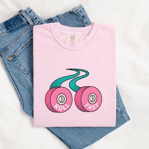 May include: A light pink t-shirt with a graphic of two pink roller skate wheels connected by a teal wavy line. The words "ROLL FAST" are printed on the wheels. The shirt is laid on top of a pair of blue jeans.
