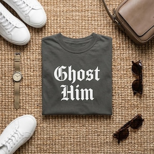 May include: A dark gray t-shirt featuring the white text "Ghost Him" in a gothic font, surrounded by a pair of white sneakers, a brown leather shoulder bag, a wristwatch with a tan strap, and two pairs of dark brown tortoiseshell sunglasses.