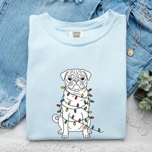 May include: A light blue Comfort Colors t-shirt featuring a white pug dog illustration wrapped in colorful Christmas lights. The shirt is folded on a denim surface, with a small succulent plant and jewelry visible.