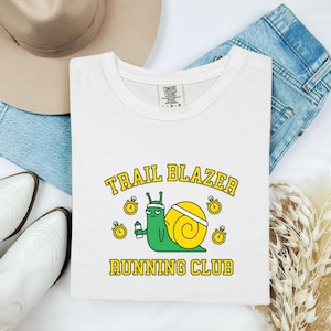 Könnte beinhalten: Weißes T-Shirt mit einer Grafik einer grünen Schnecke, die eine Wasserflasche hält, mit einem gelben Gehäuse und den Worten "TRAIL BLAZER RUNNING CLUB" in Gelb und Grün. Das Design enthält gelbe Uhren.