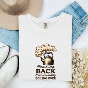 May include: A white short-sleeve t-shirt featuring a graphic of a metallic kettle emitting a large cloud of steam. Below the kettle, the text reads: Please step BACK I am currently BOILING OVER. The shirt has a Comfort Colors brand neck label.