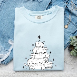 May include: A light blue t-shirt with a graphic of a Christmas tree made of white cats. The shirt has the Comfort Colors brand tag. The design includes a star at the top and smaller stars around the tree.