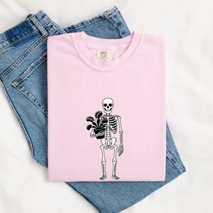 May include: A light pink t-shirt featuring a black line art graphic of a human skeleton holding a potted Venus flytrap plant. The shirt has a crew neckline and a Comfort Colors brand label printed on the inside of the collar area.