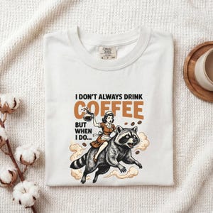 Puede incluir: Una camiseta blanca con el gráfico de una persona con uniforme marrón montada sobre un mapache mientras sostiene una cafetera. El texto dice: I DON'T ALWAYS DRINK COFFEE BUT WHEN I DO... La camiseta tiene una etiqueta Comfort Colors en el cuello.