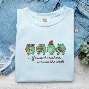 May include: Light blue t-shirt with the text "caffeinated teachers survive the week." The shirt features a graphic of four cartoon frogs, each holding items like a coffee cup, books, and an apple. The shirt is folded on a denim surface.
