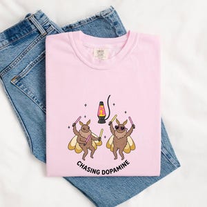 May include: A light pink t-shirt featuring a graphic of two brown moths with yellow wings, wearing sunglasses and holding glow sticks. A lava lamp is positioned between them, and the text "CHASING DOPAMINE" is printed in black below the illustration.