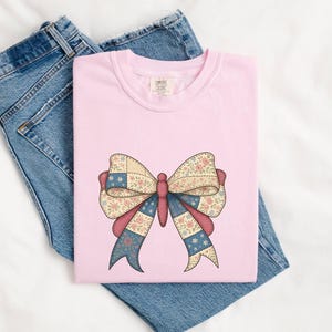 May include: A pale pink t-shirt with a large bow design. The bow features a patchwork pattern with floral and star details in pink, blue, and cream. The shirt is placed on top of a pair of blue denim jeans.