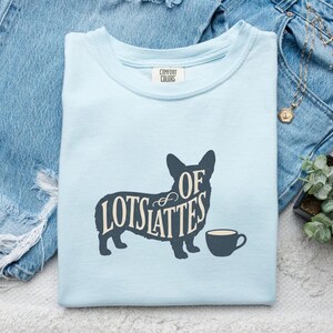 Puede incluir: Una camiseta azul claro con el texto "Lots of Lattes" en fuente azul oscuro, diseñado con la forma de un perro corgi. Una pequeña taza de café azul oscuro está al lado del perro. La camiseta está doblada sobre unos vaqueros azules.