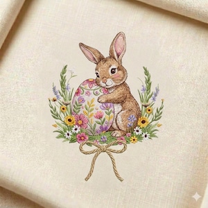 Peut inclure: Broderie représentant un lapin brun câlinant un œuf de Pâques décoré. L'œuf est orné de motifs floraux roses, violets et jaunes. Le lapin et l'œuf sont entourés d'une couronne de fleurs et de verdure, avec un nœud noué en bas.