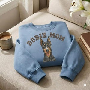 Può includere: Una felpa azzurra con le parole "DOBIE MOM" ricamate in lettere marroni sopra un'illustrazione dettagliata di un Dobermann Pinscher. Il muso del cane è nero, marrone e beige. La felpa ha polsini a coste e girocollo.