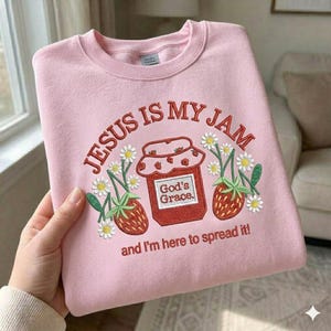 Può includere: Felpa rosa chiaro girocollo con la scritta ricamata "JESUS IS MY JAM" sopra un barattolo di marmellata e fragole. Il barattolo riporta la scritta "God's Grace", con la frase "and I'm here to spread it!" sotto.