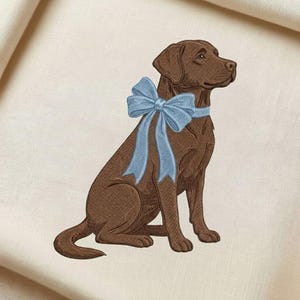 May include: An embroidered image of a brown Labrador Retriever dog wearing a light blue bow. The dog is sitting upright, facing right, with its head turned slightly. The background is a cream-colored fabric.