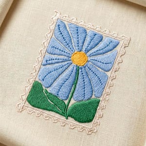 May include: Embroidered blue flower design on cream fabric. The flower has a yellow centre and green leaves, framed by a decorative border. The fabric is folded, suggesting a textile item.
