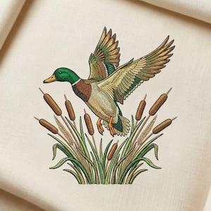 May include: Embroidered image of a mallard duck in flight, featuring a green head, brown chest, and patterned wings. The duck is depicted above cattails and reeds, set against a cream-coloured background.