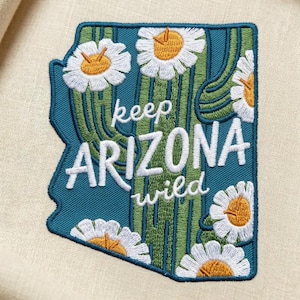 Puede incluir: Parche bordado con la forma de Arizona, con fondo turquesa, diseño de cactus y margaritas blancas. El texto "keep ARIZONA wild" está bordado en blanco.
